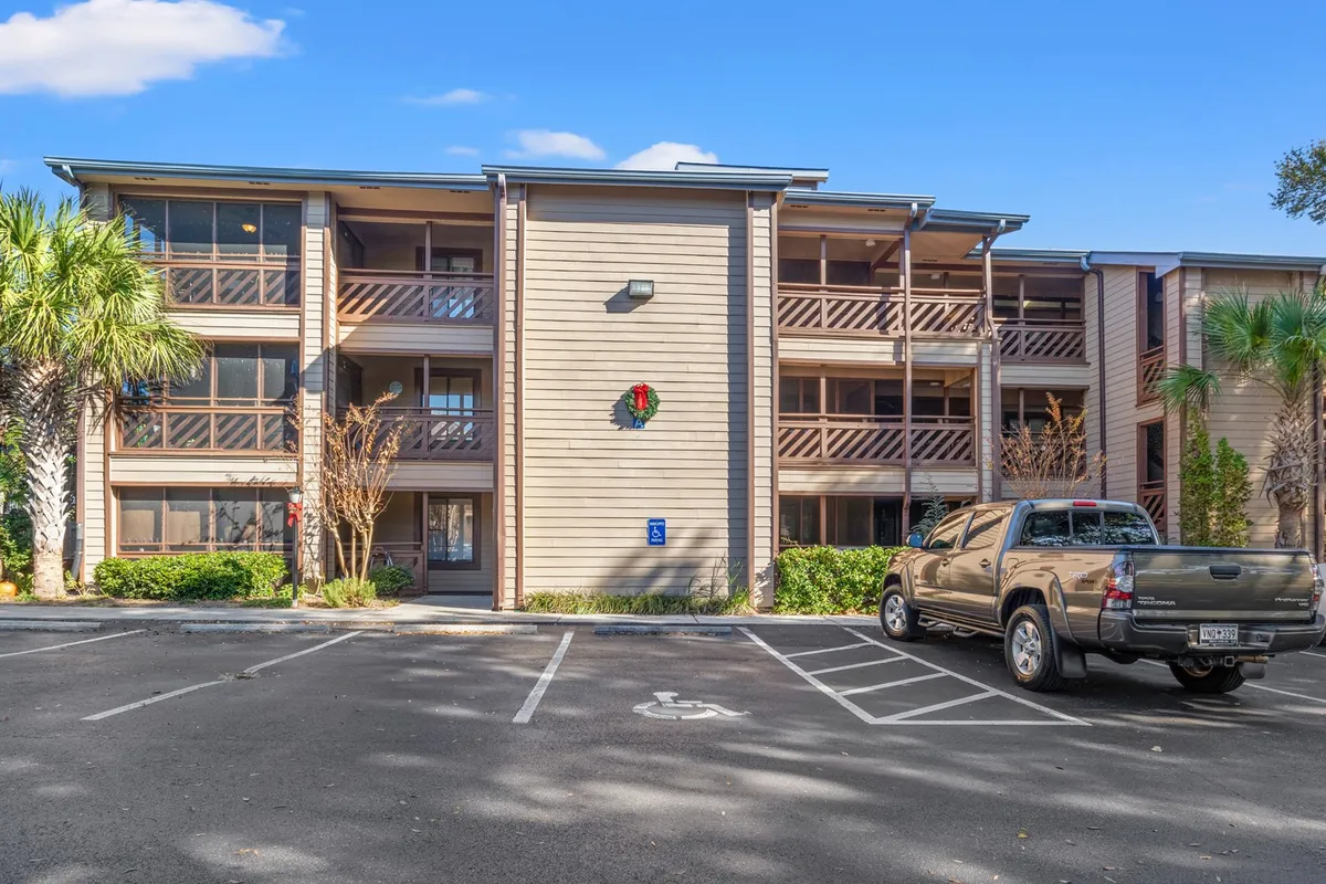 The image depicts the exterior of a welcoming residential building, featuring a balcony with a wooden railing adorned with a decorative wreath. Tall palm trees frame the scene, and a parking area with a truck sits in front of the building. The ambiance is homey and inviting, suggesting a tranquil setting ideal for long-term stays.