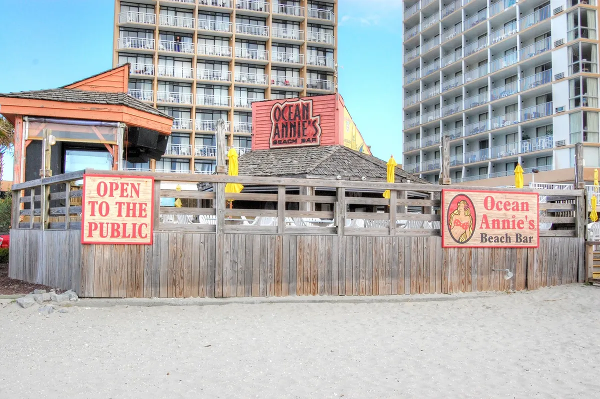 This image features the Ocean Annie's Beach Bar sign with yellow umbrellas dotting a sunlit wooden deck. The space is lively with groups of friends socializing, enjoying drinks and food, and soaking up beach views. The bar’s inviting, public setting and proximity to the ocean create an energetic, group-friendly ambiance perfect for gatherings and nightlife.