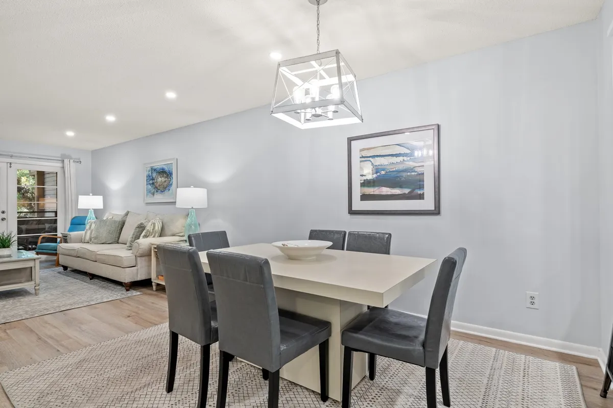 This image features a dining area with a table set beneath modern chandeliers, a sofa visible in the background, and artistic decor on the walls. The scene suggests a seamless connection between the dining and living spaces, reinforcing the open-concept design.