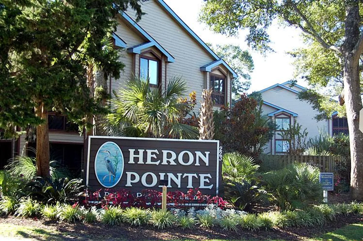This image features welcoming signage for Heron Pointe, surrounded by lush landscaping and the architectural details of the property buildings. The scene exudes tranquility, with green gardens and mature trees offering a serene introduction to the community.