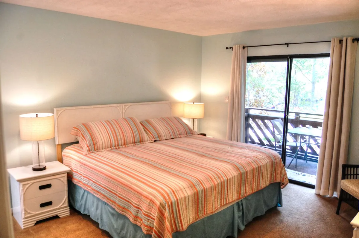A bright bedroom featuring a king-sized bed with striped bedding, nightstands with lamps on either side, and a large sliding glass door that lets in gentle natural light. The room feels airy, inviting, and perfect for restful mornings.