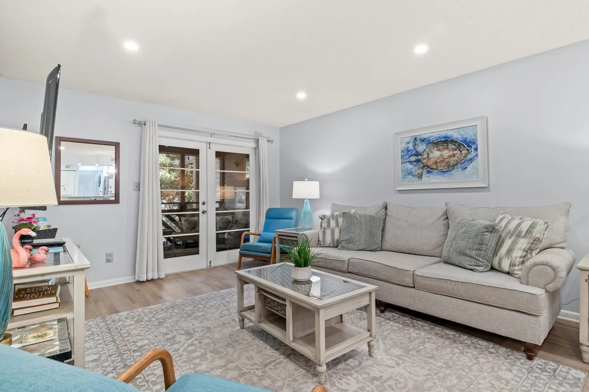 The image showcases the inviting living room of a Myrtle Beach condo, featuring a comfortable sofa, modern coffee table, and tasteful wall art. Large sliding glass doors bathe the space in natural light and open to tranquil nature views, hinting at sunrise vistas and a seamless connection to the outdoors.