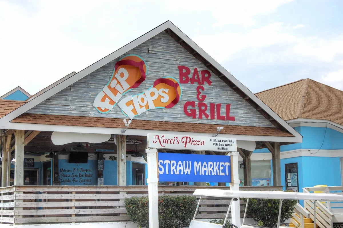 Lively exterior signage for Flip Flops Bar & Grill, Nucci's Pizza, and the Straw Market creates a vibrant scene reflecting the nearby dining and entertainment options. The mood is casual and inviting, capturing the essence of local outings.