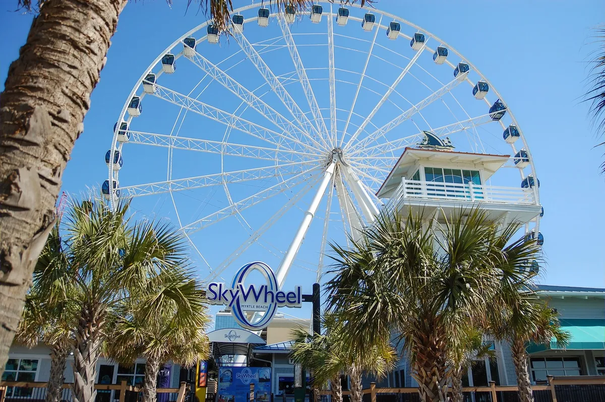 A lively shot of the SkyWheel with palm trees framing the foreground and the ticket booth visible, suggesting a vibrant boardwalk atmosphere. Visitors are seen enjoying the ferris wheel ride and the surrounding beach attractions.